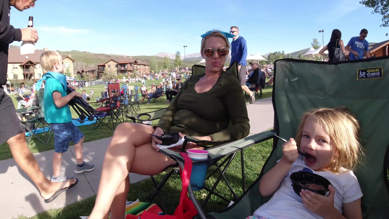 A close chair-level view shows a woman and child in the foreground, a boy standing near the path, and a large audience spread across the grass in Winter Park, Colorado.