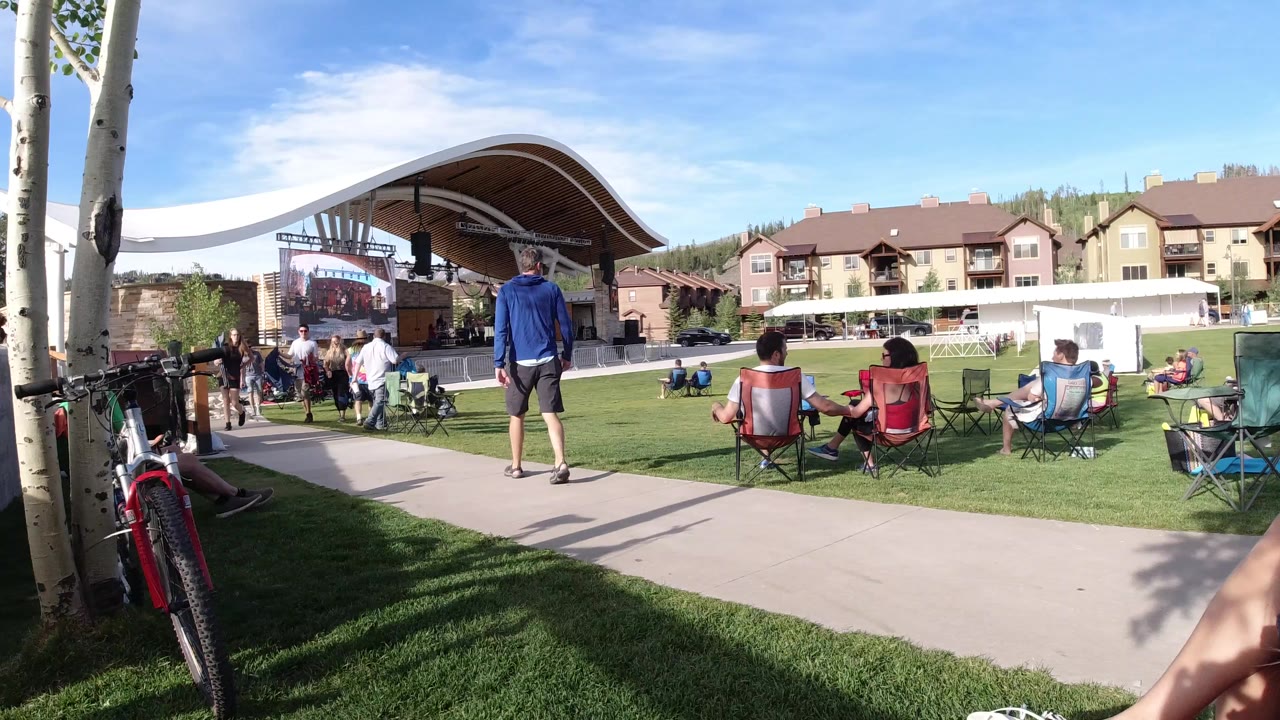 Wide view in Winter Park, Colorado of bicycles beside aspen trunks, seated spectators on the grass, people near a covered stage, and lodge-style buildings in bright sun.