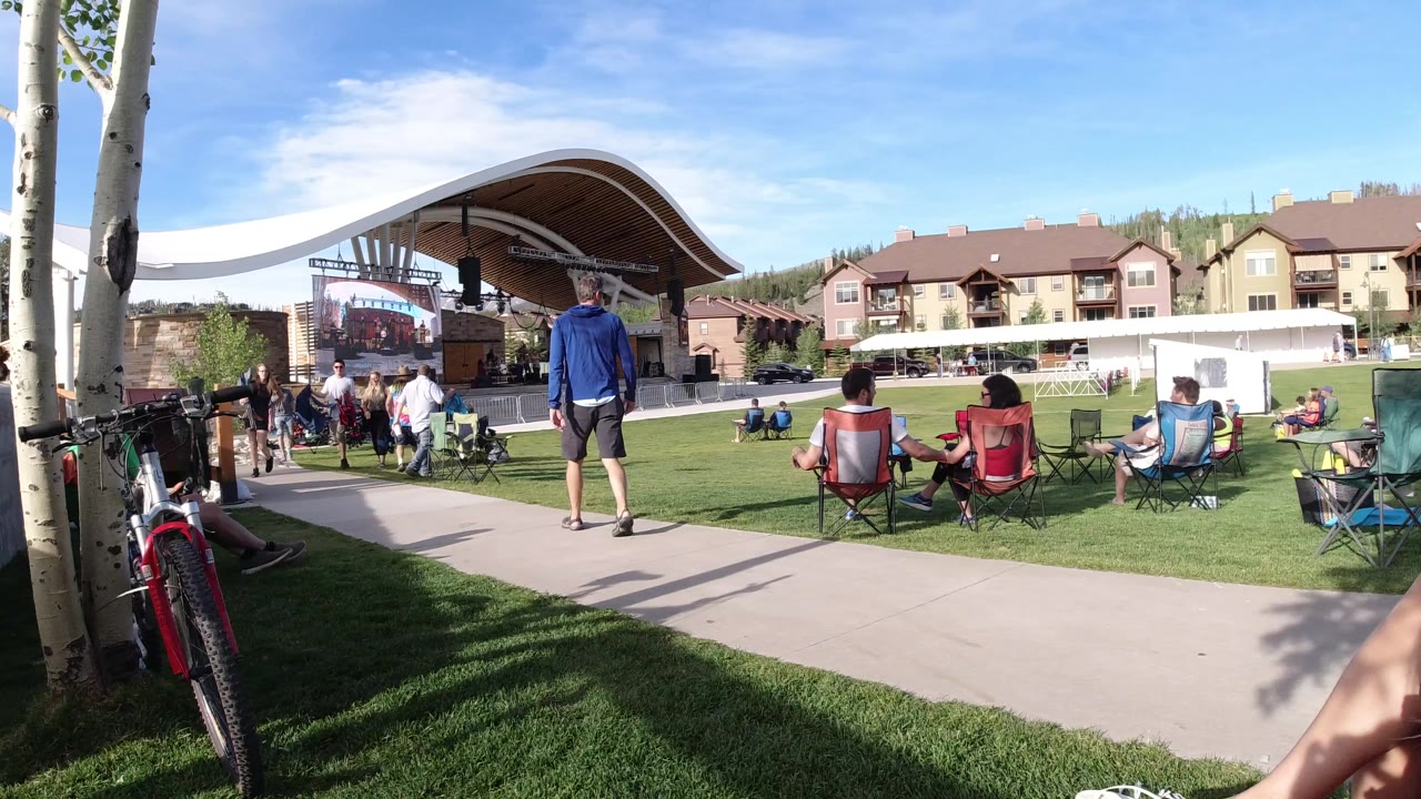 A man in a blue top stands on the paved path in Winter Park, Colorado while spectators sit in folding chairs facing a covered outdoor stage.
