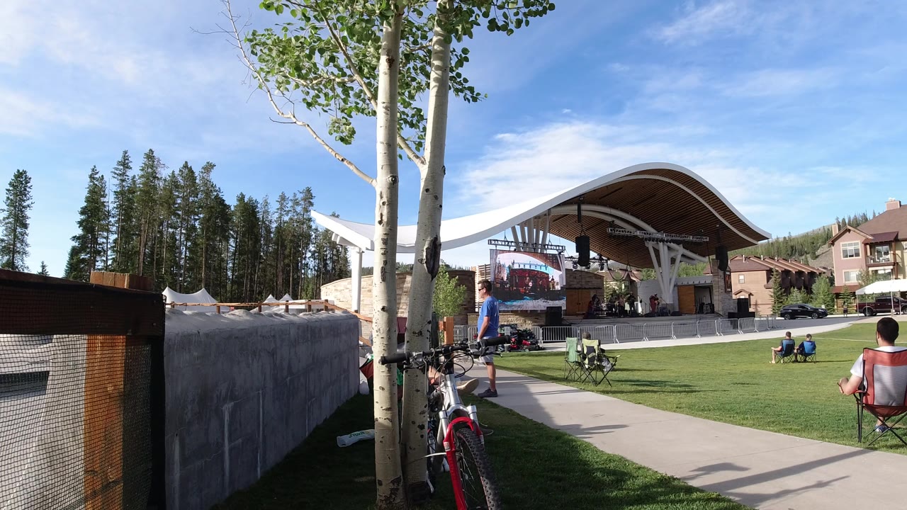 Wide lawn view with a covered stage, trees, bicycle, and seated people.