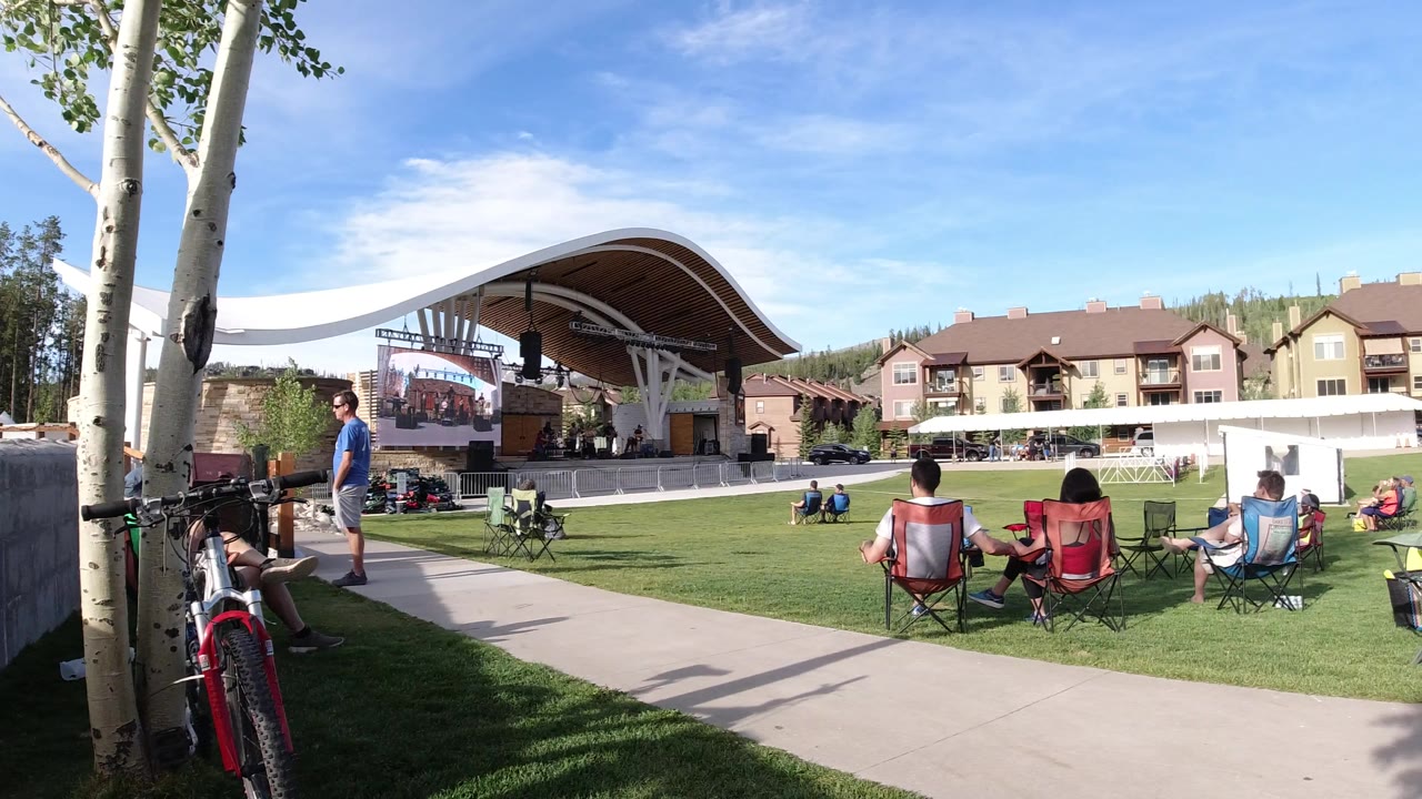 A man stands near the paved path while seated spectators look toward the covered stage at the Winter Park, Colorado outdoor gathering.