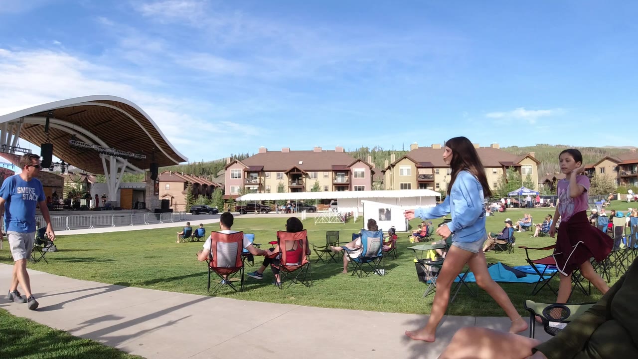 Wide ground-level view in Winter Park, Colorado, with people on folding chairs across a lawn, two women walking on a paved path, a curved stage canopy, low buildings, hills, and blue sky.