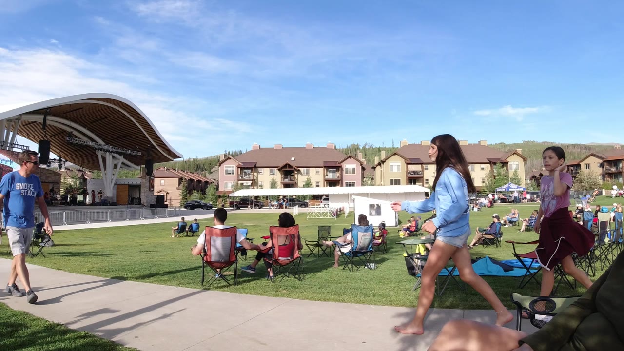 Winter Park, Colorado lawn scene with a man in a blue shirt and two women walking near seated spectators, backed by a curved stage canopy, resort-style buildings, hills, and open sky.