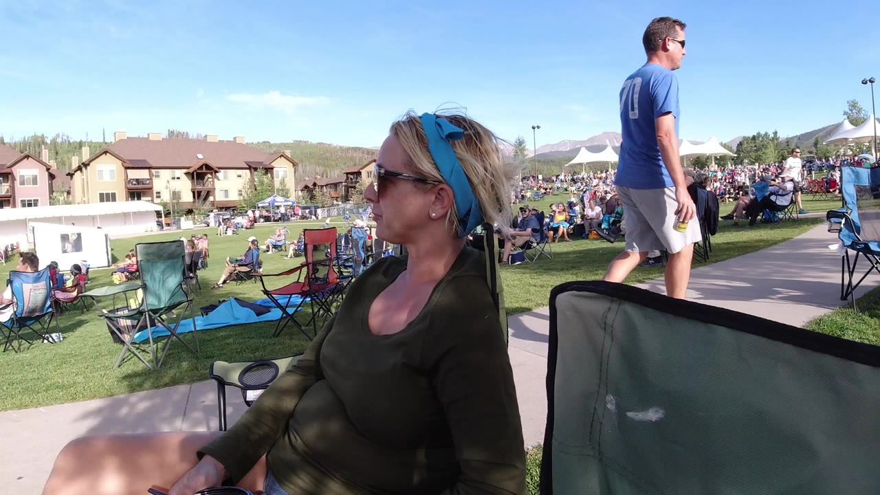 Woman in sunglasses seated near a crowd on a sunny lawn.