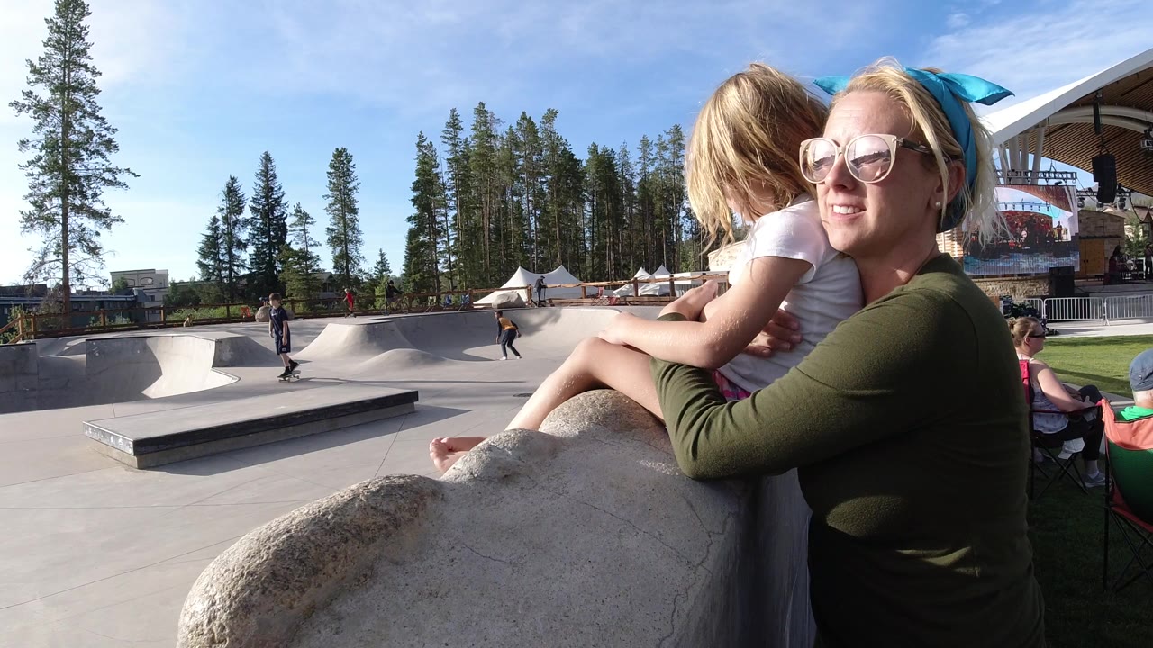 Woman in sunglasses holding a child beside an outdoor skate park.