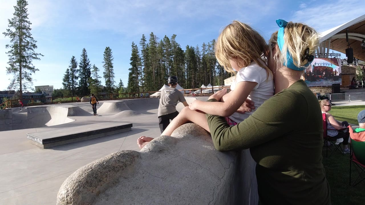 Mother holding a child beside a concrete skate park with riders in the distance.