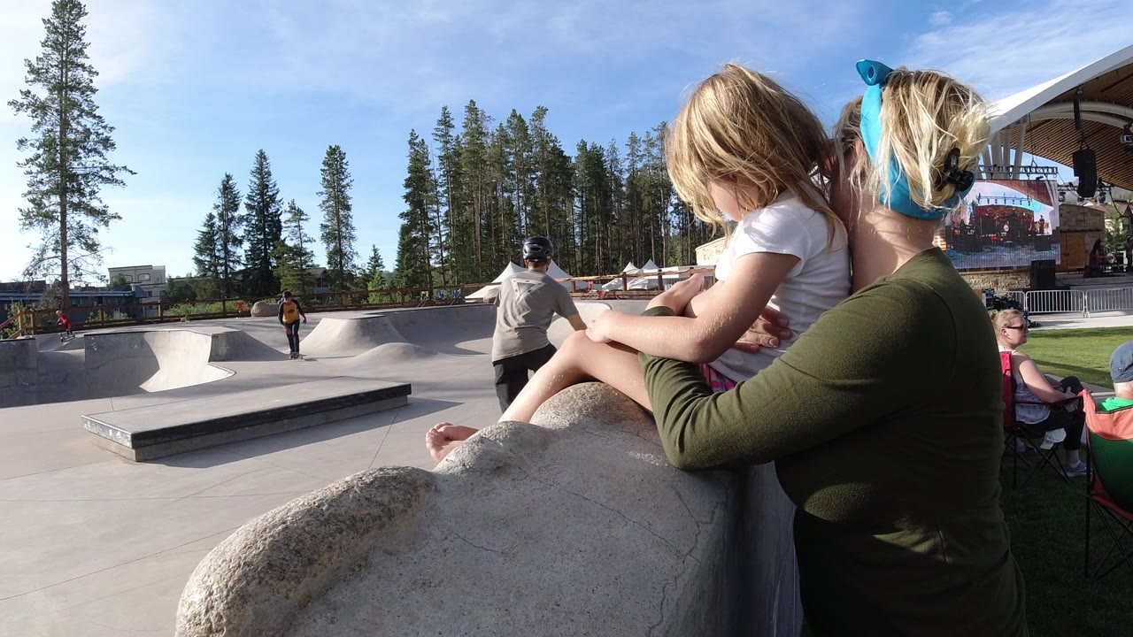 The mother and child remain in the right foreground in Winter Park, Colorado, as a skateboarder crosses the open concrete park near low ramps and rails.