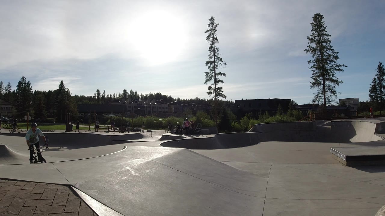 Wide view of a Winter Park, Colorado skate park with a cyclist entering from the left, concrete bowls in the midground, tall trees and buildings in the background, and strong sun glare.