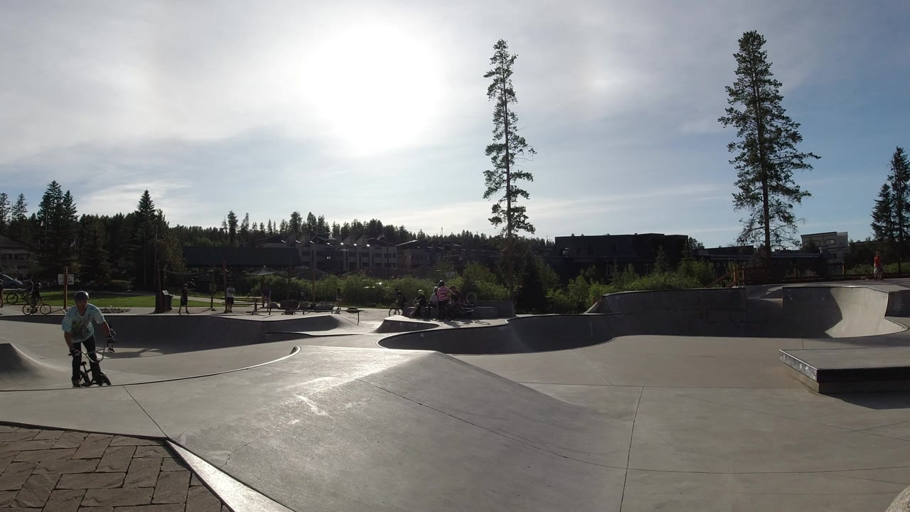 A bicycle rider moves closer to the foreground edge of the Winter Park, Colorado skate park, with curved concrete bowls, a ramp platform, trees, and backlit buildings behind.