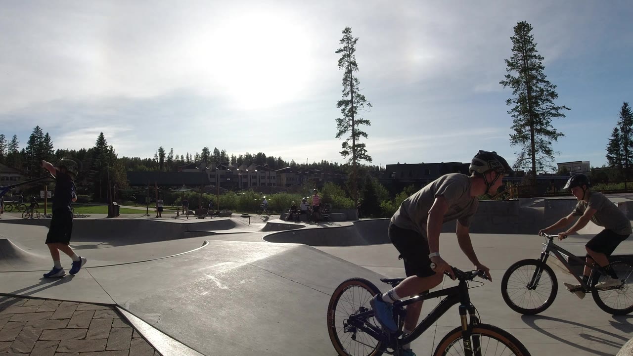Children riding bikes and a scooter at a concrete skate park.