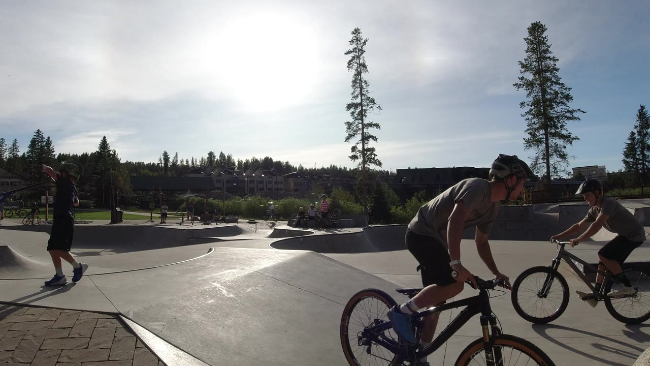 Two riders on bicycles and a child holding a scooter cross the frame at the Winter Park skate park, with concrete bowls, trees, and strong backlight visible.
