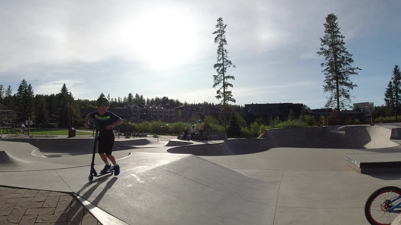 A boy stands with a scooter in the foreground of a sunny concrete skate park in Winter Park, Colorado, with ramps, trees, and buildings behind him.