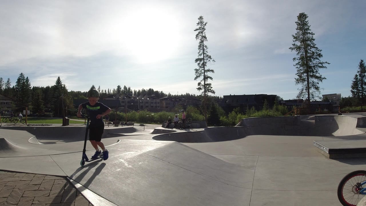 A child on a scooter stands near the foreground while the concrete ramps and tree-lined background remain visible in the Winter Park skate park.