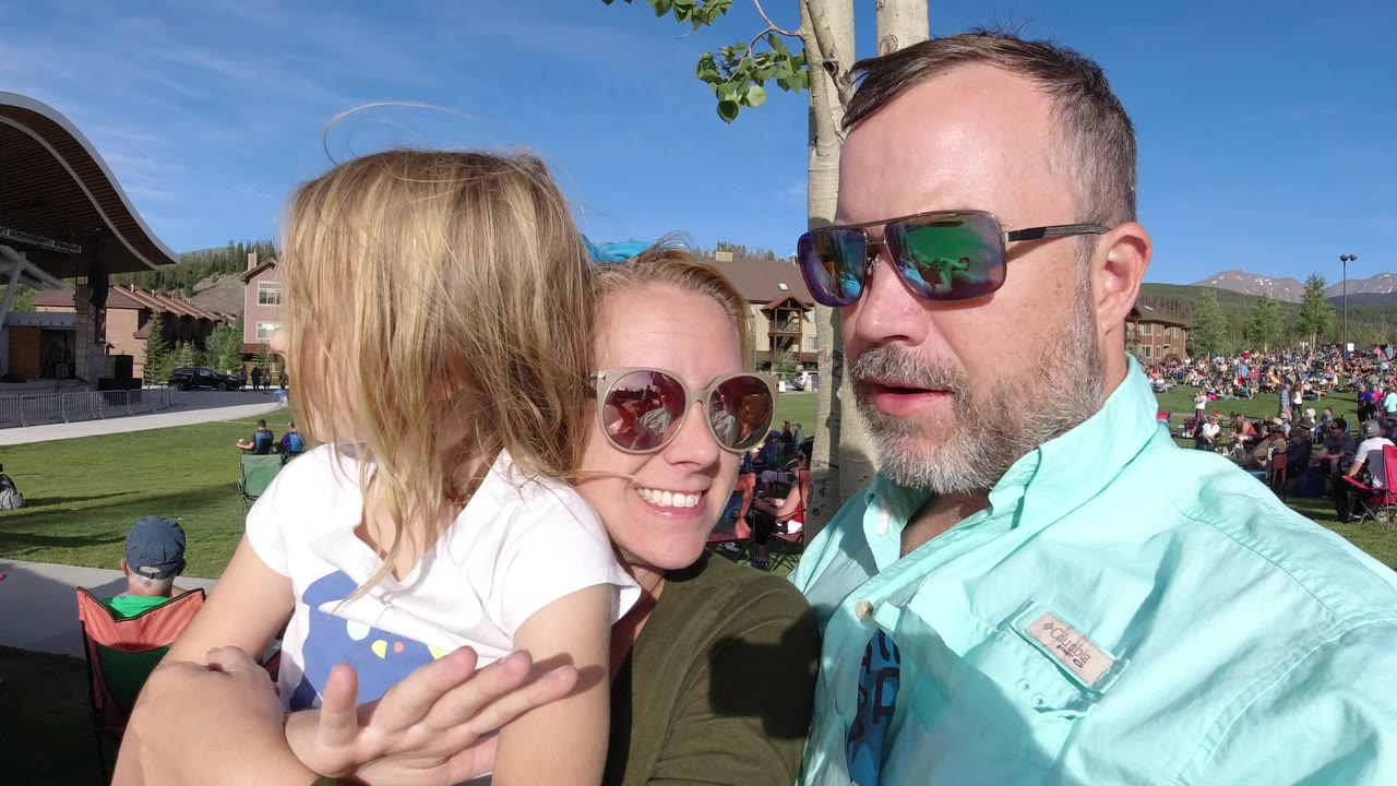 At 0:01 in Winter Park, Colorado, the woman smiles from behind the child while the man looks near the camera, with the bright lawn event and seated crowd stretching across the background.