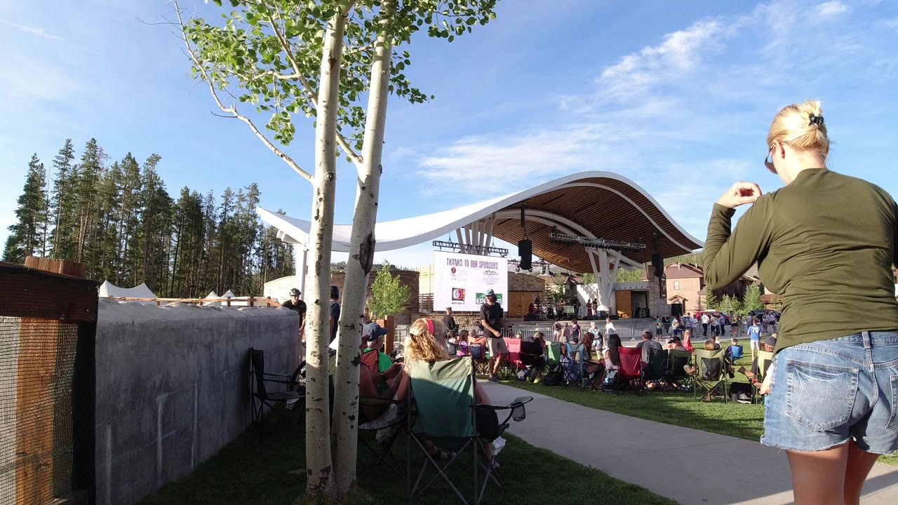The Winter Park, Colorado concert scene continues with the woman closer to the center foreground, rows of folding chairs on the grass, and a large white sign hanging at the stage.