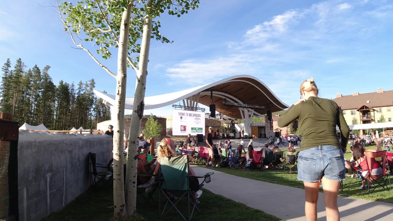 A wide frame in Winter Park, Colorado shows the woman moving toward the right edge, spectators seated across the grass, a curved-roof stage, and chalet-style buildings in the background.
