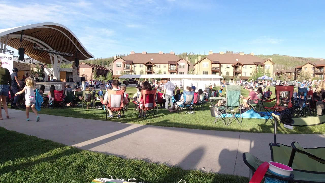 Wide view in Winter Park, Colorado of people seated in folding chairs on grass facing a curved outdoor stage, with a paved walkway and hillside buildings behind them.