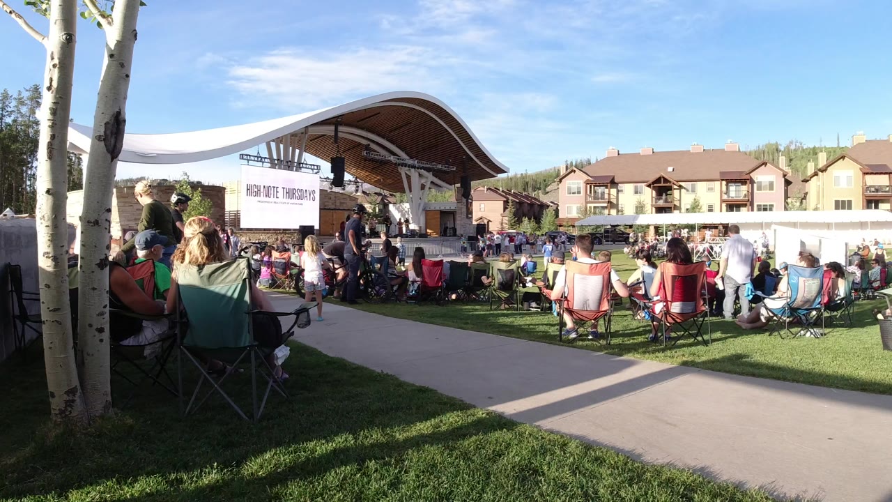 Wide lawn-level view in Winter Park, Colorado, with people in folding chairs facing a covered outdoor stage, pale tree trunks at left, and buildings behind the crowd.