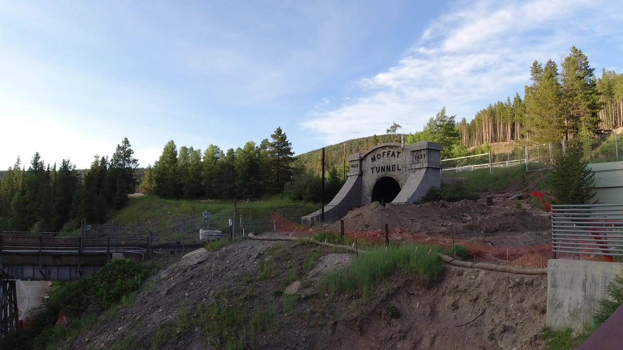 Concrete tunnel portal on a green hillside with trees and construction fencing.