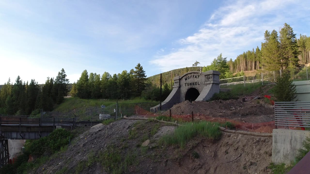 Winter Park frame with the Moffat Tunnel entrance centered near the hillside, a metal railing in the foreground, orange fencing across the dirt, and no visible people.