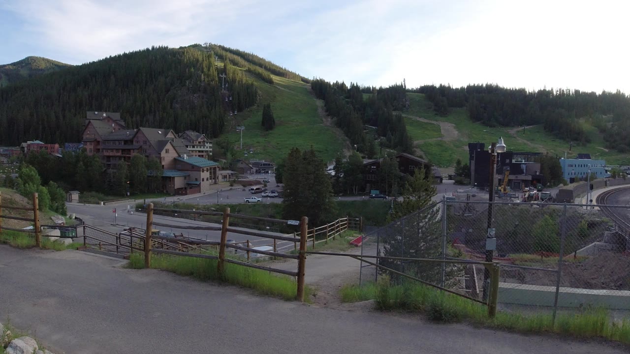Wide hilltop view of buildings, road, fences, green slopes, and forested mountain.