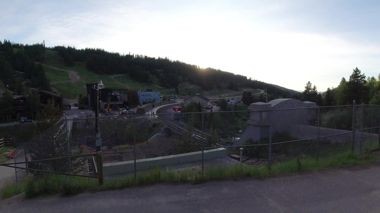 Wide view in Winter Park, Colorado of a paved path, chain-link fence, concrete structures, industrial buildings, forested hillside, low sun, and very small distant people near the work area.