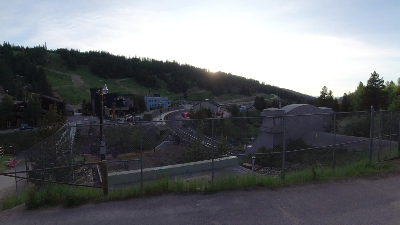 Winter Park, Colorado frame with the fence line centered, a concrete utility structure to the right, industrial buildings to the left, wooded hills behind, and tiny distant people near the site.