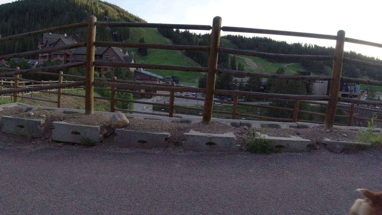 A wooden rail fence in Winter Park, Colorado overlooks a road, lodge-style buildings, green hillside, and dark evergreen forest, with no visible people.