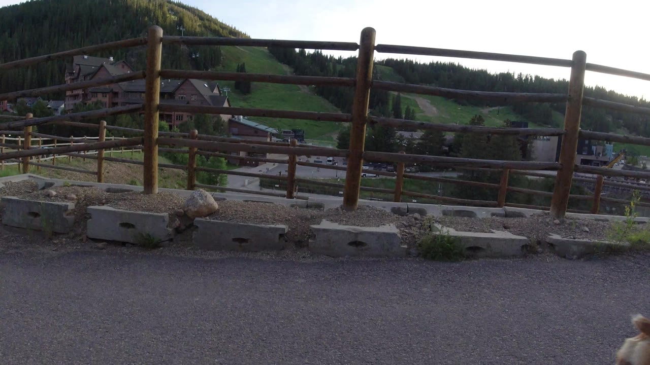 The second frame keeps the wooden fence and concrete barriers prominent above a Winter Park, Colorado road, buildings, hillside grass, and tree line, with no visible people.
