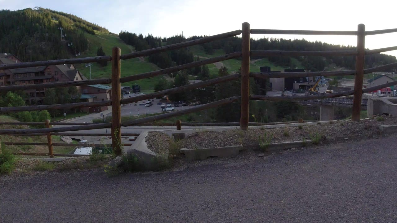 Opening frame of a wooden railing and gravel overlook above roads, parked vehicles, chalet-style buildings, green hillside, and pine forest in Winter Park, Colorado, with no visible people.