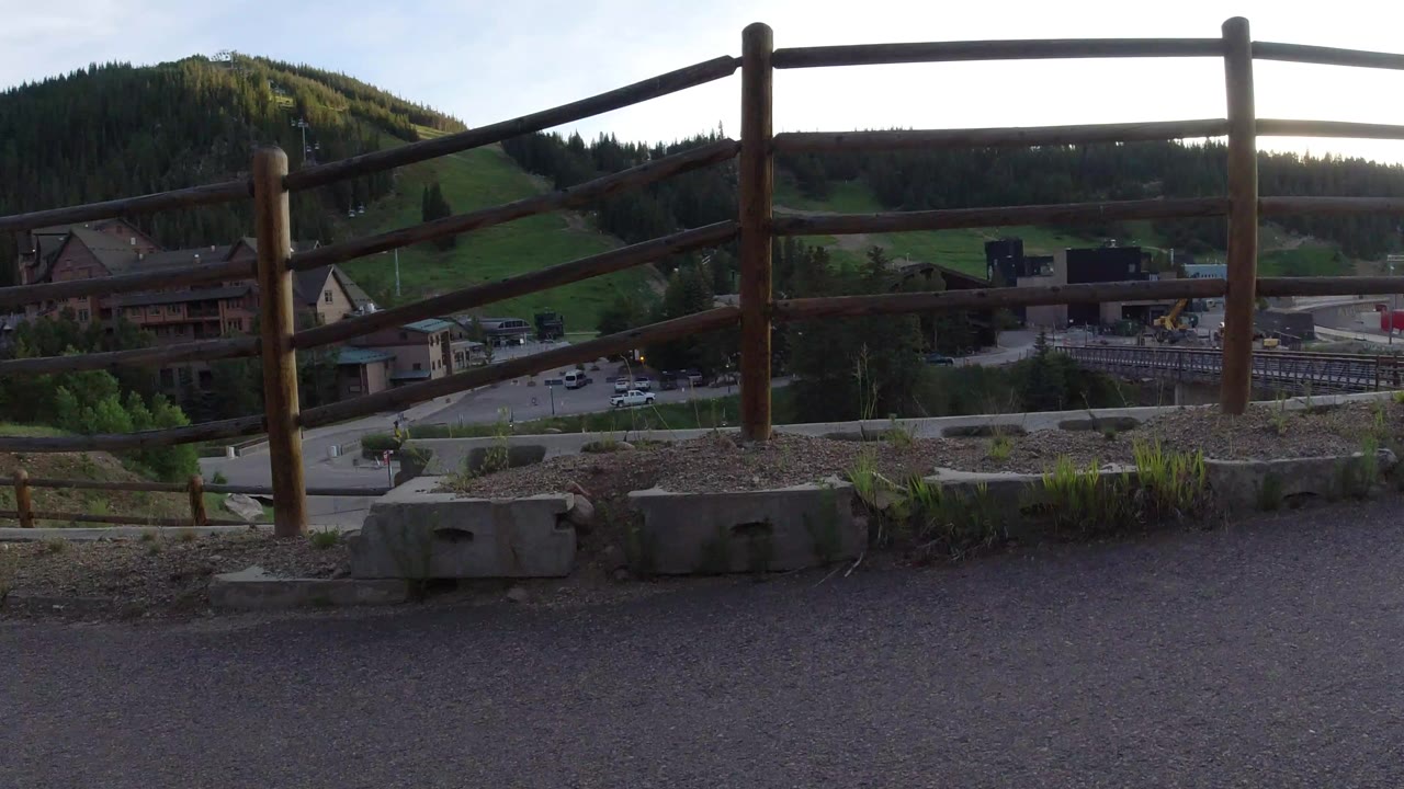 Opening frame of a Winter Park, Colorado overlook with a timber fence in front, resort-area buildings and roads below, forested hills beyond, and no visible people.