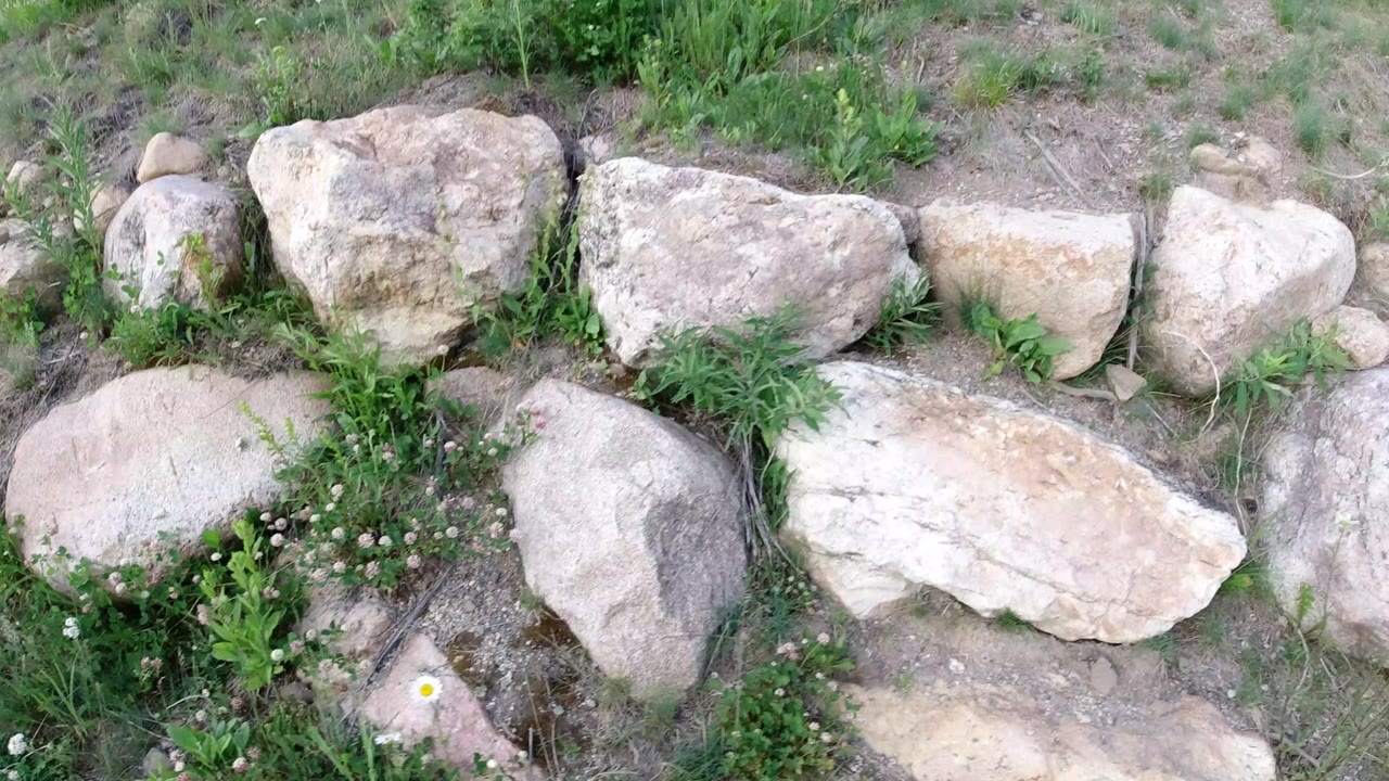 A similar downward view shows rounded boulders, weeds, clover-like white flowers, and exposed dirt along the Winter Park rock edge, with no visible people.