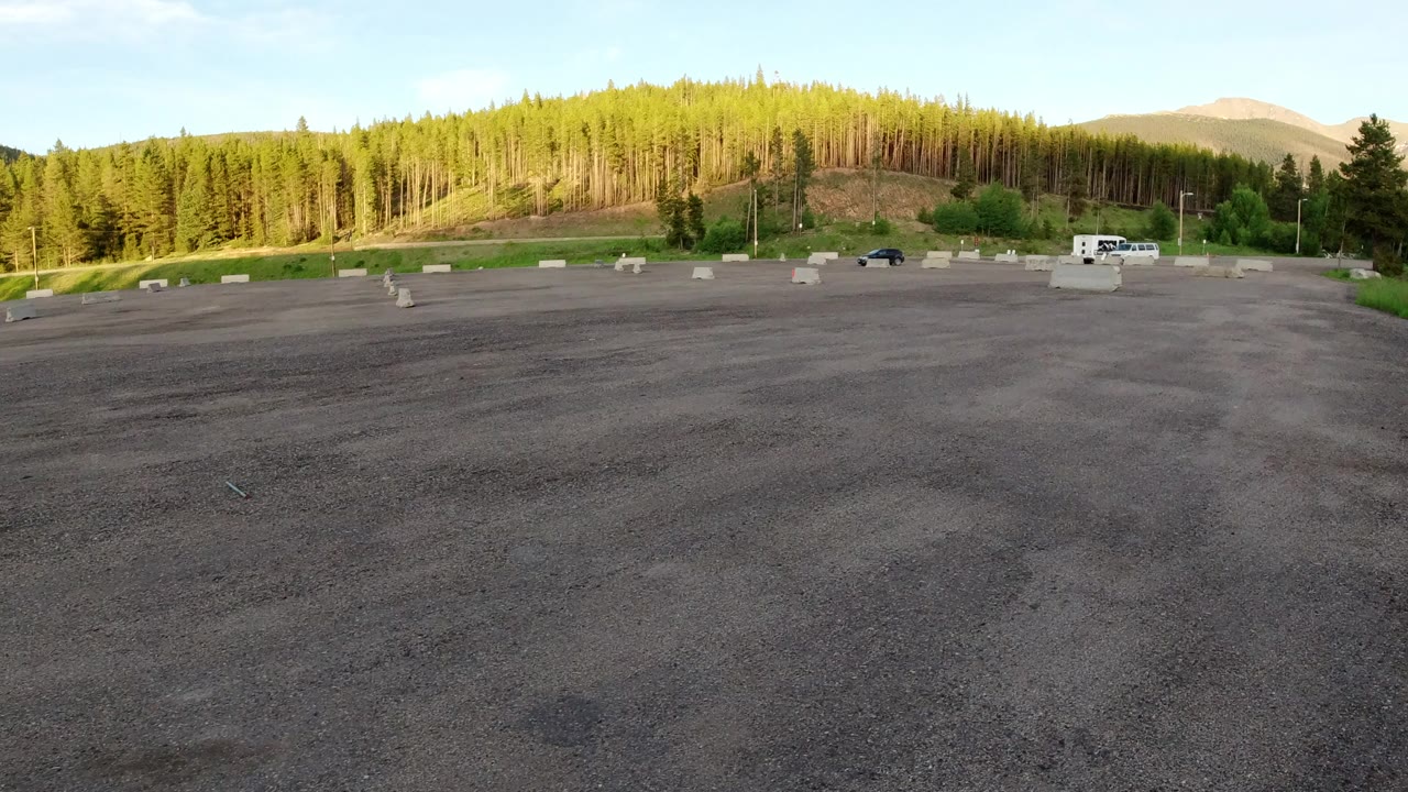 Wide view of a paved lot in Winter Park, Colorado, with concrete barriers across the middle distance, sunlit evergreen hills, small vehicles, and no visible people.