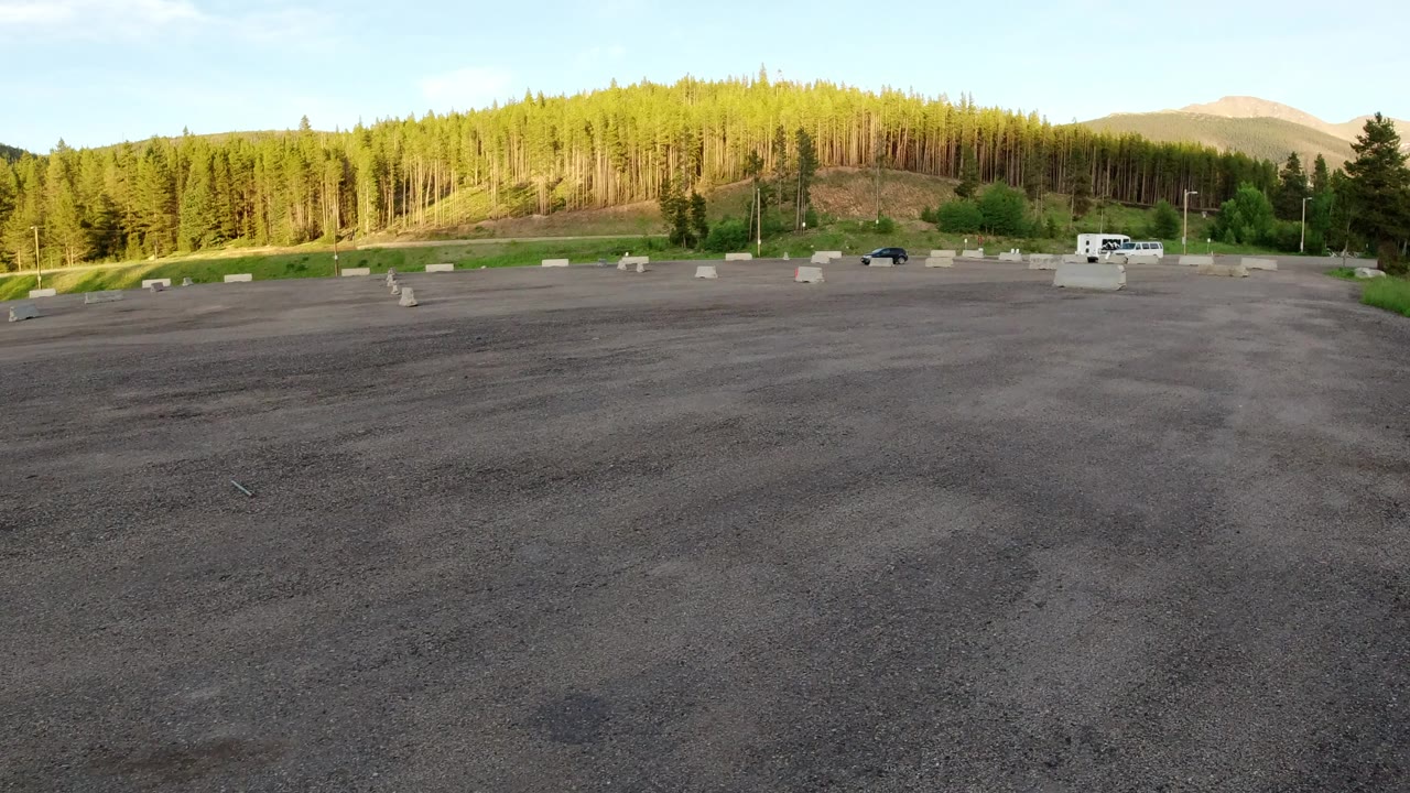 A broad asphalt foreground leads to scattered concrete parking barriers and a green hillside in Winter Park, Colorado, with a dark vehicle near the road and no visible people.