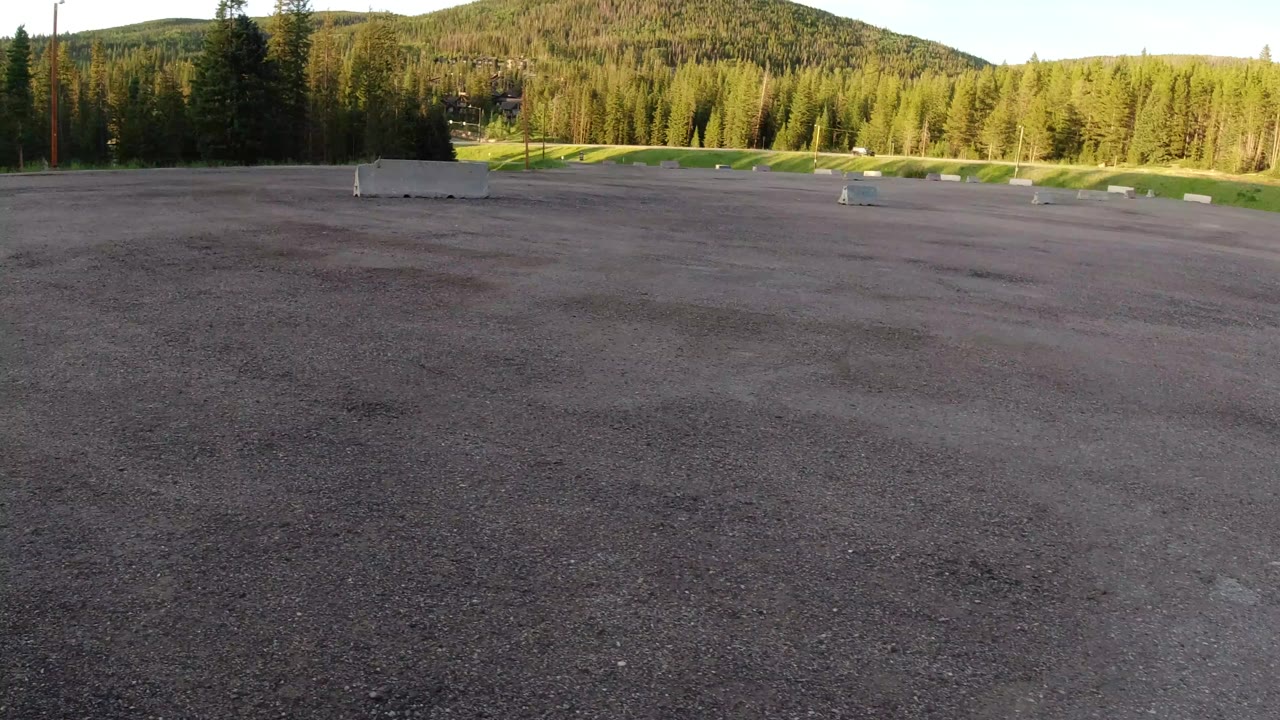 Wide view of an empty gray lot in Winter Park, Colorado, with concrete barriers, green grass, evergreen trees, and a low hillside in warm light, no visible people.