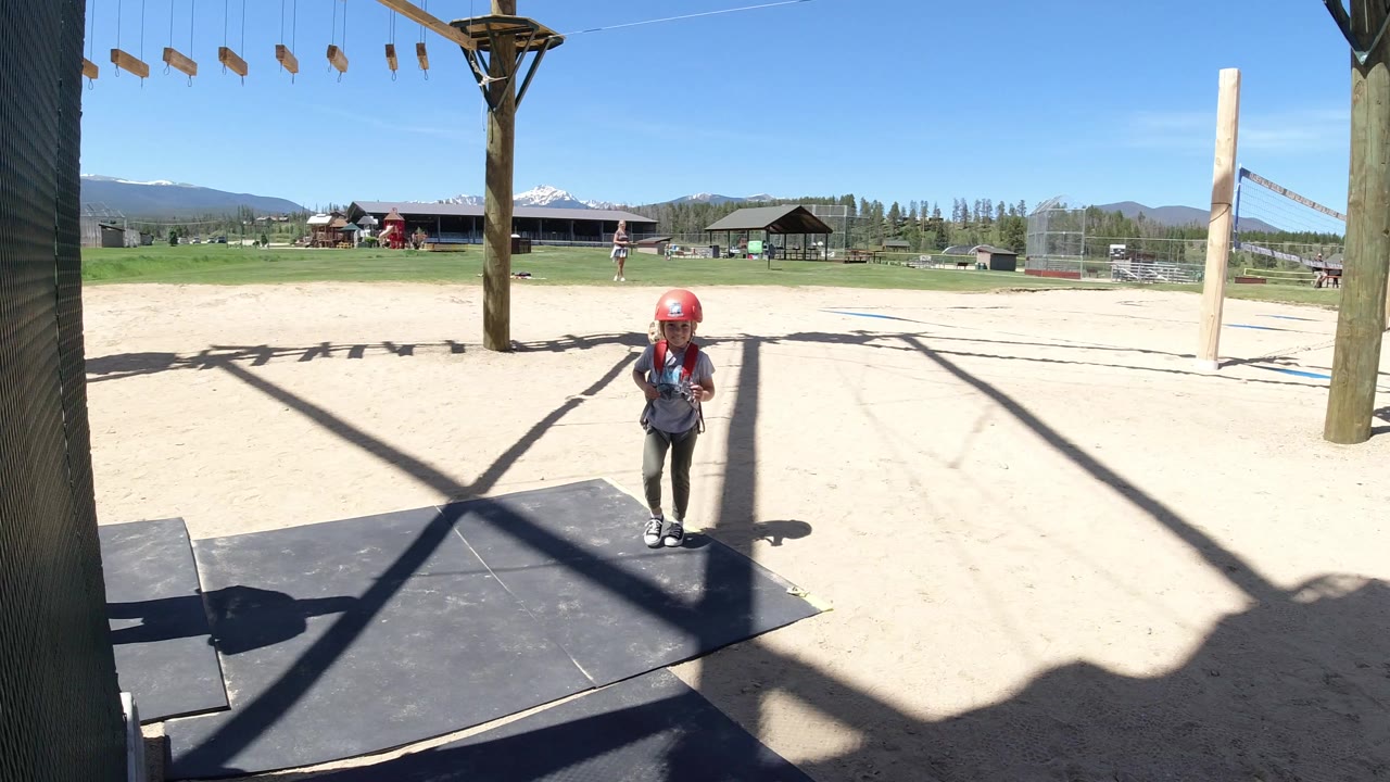 The girl remains centered in the Winter Park activity area, standing on the mat while overhead ropes, sandy ground, buildings, and distant mountains fill the background.