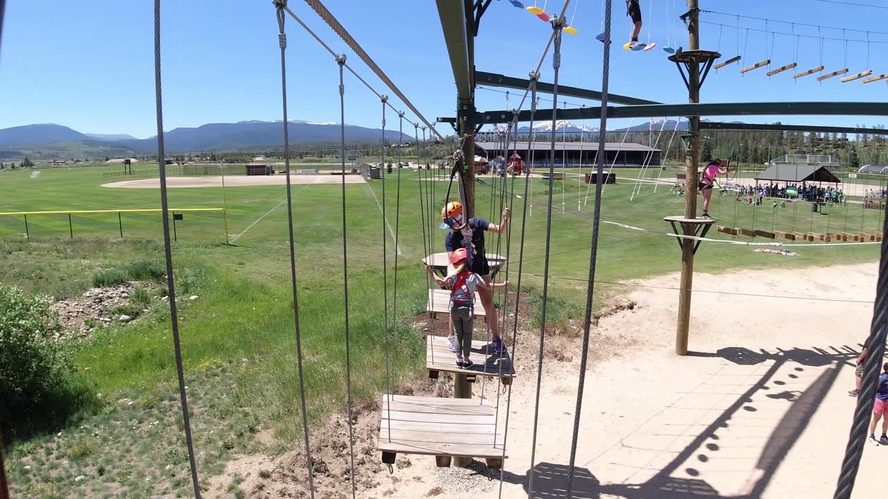 Two helmeted participants remain on the wooden platform while another person is visible farther along the Winter Park, Colorado ropes course above the grassy recreation area.