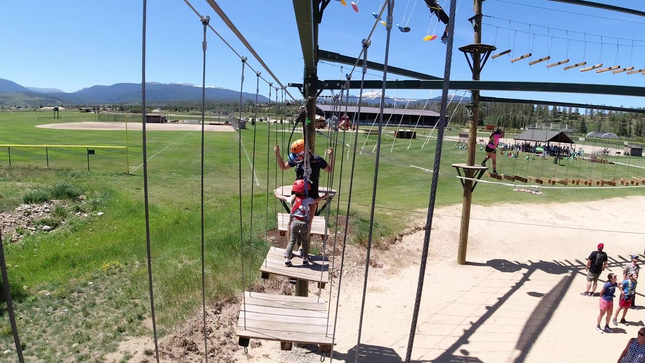 People in helmets stand on wooden ropes-course platforms in Winter Park, Colorado, with vertical cables in the foreground, green fields, and distant mountains behind them.
