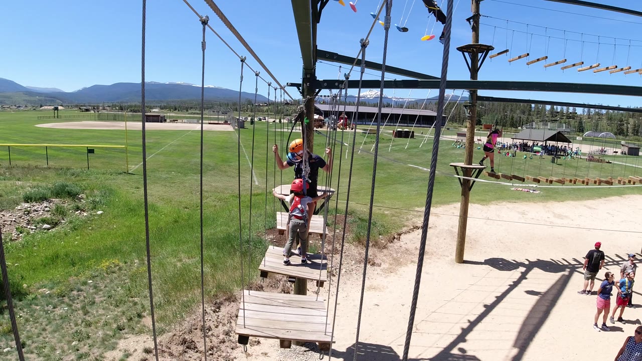 A helmeted participant stands on a small wooden platform while other people wait below, with the Winter Park, Colorado field and mountain horizon visible beyond the ropes course.