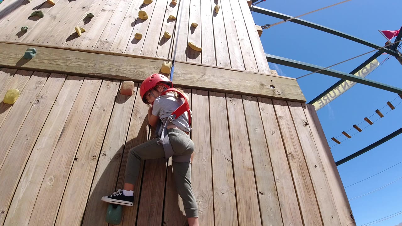 Child in helmet and harness climbing a wooden wall.