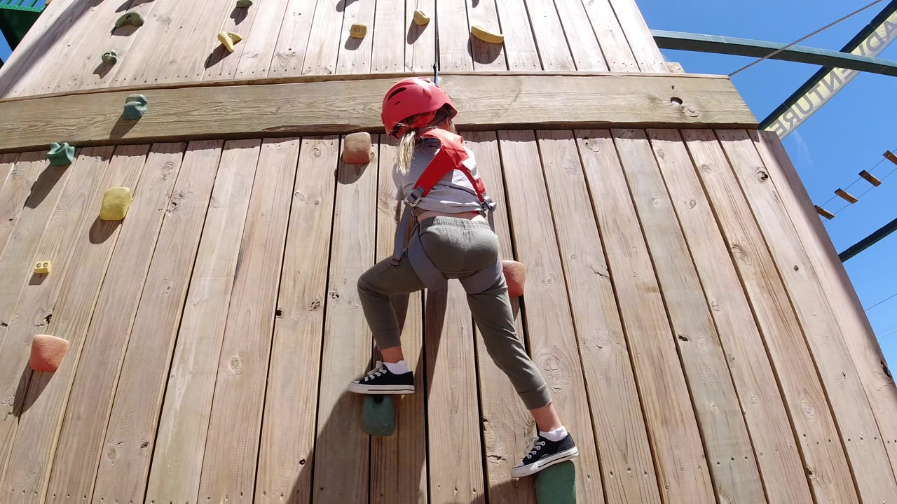Girl in a red helmet climbs a wooden wall with colored holds.