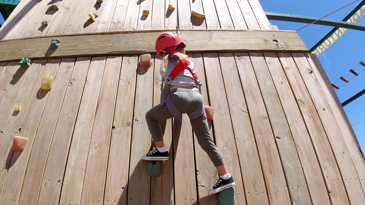 At 0:01, the child climber remains centered on the wooden wall in Winter Park, Colorado, with one knee raised and colored holds scattered around her.
