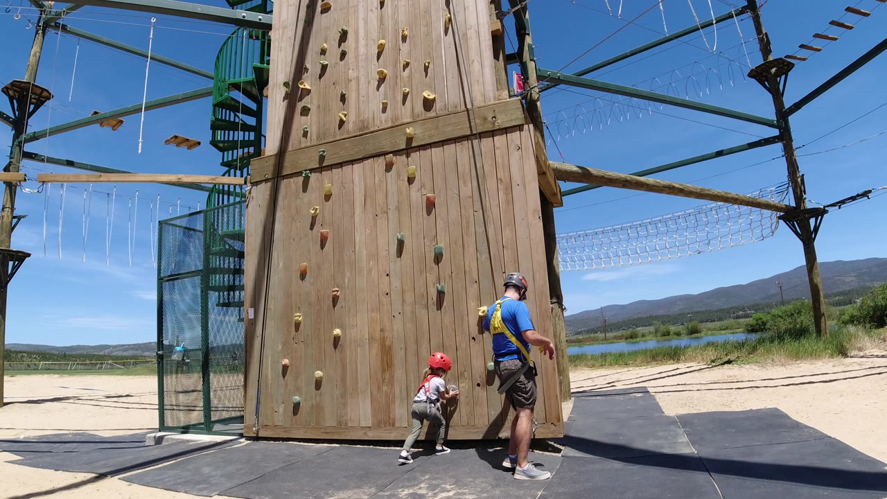 In Winter Park, Colorado, the child waits at the lower climbing holds as the instructor steps along the padded area beside the tall wooden wall and overhead rope course structure.
