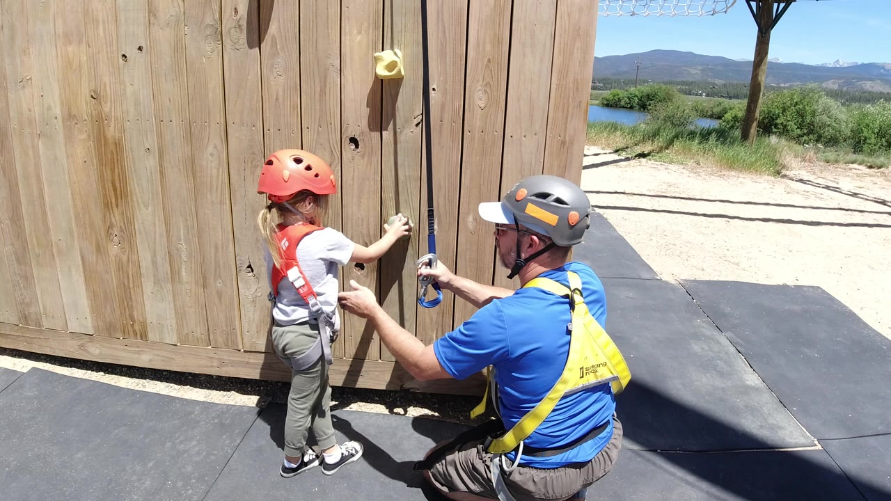 Adult man adjusts a young girl's climbing gear beside a wooden wall.