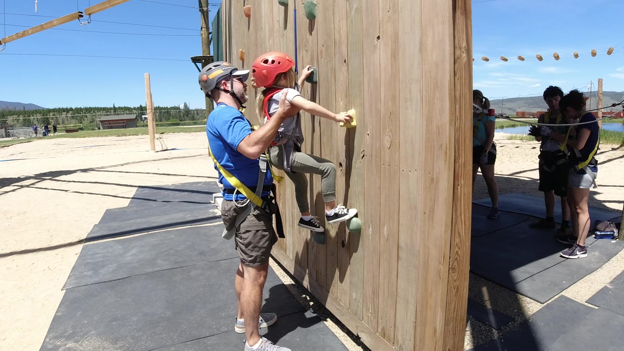 Child climbing a wooden outdoor wall with an adult supervising close by.