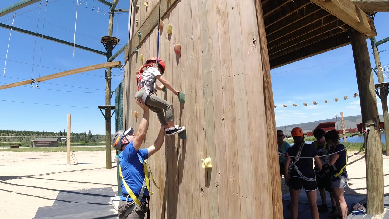 At 0:00 in Winter Park, Colorado, a helmeted girl in a harness starts up a wooden climbing wall as an adult below steadies her foot.