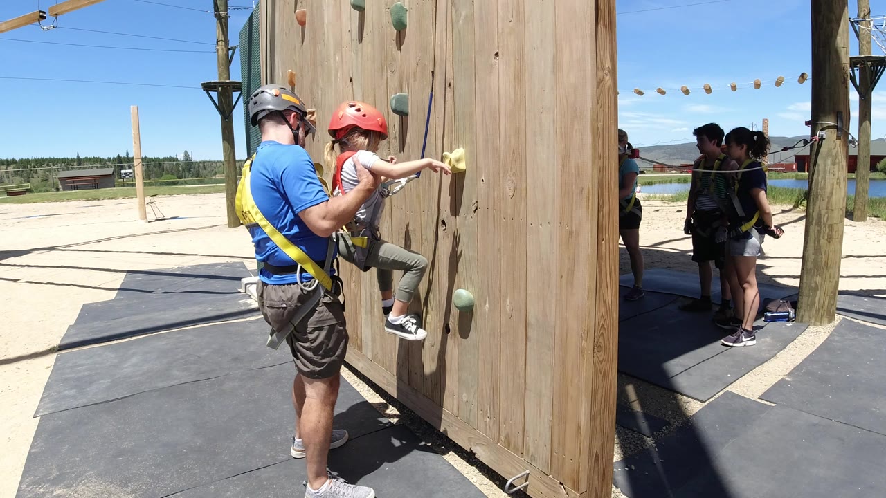 Father supports daughter on a wooden climbing wall.