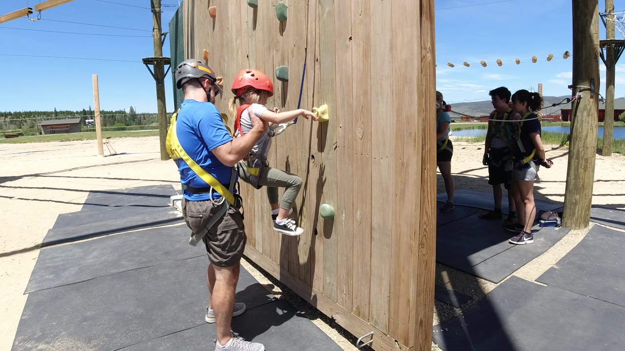 In Winter Park, Colorado, the father stands behind the child on the wooden wall while several harnessed people wait in the shaded ropes course area behind them.