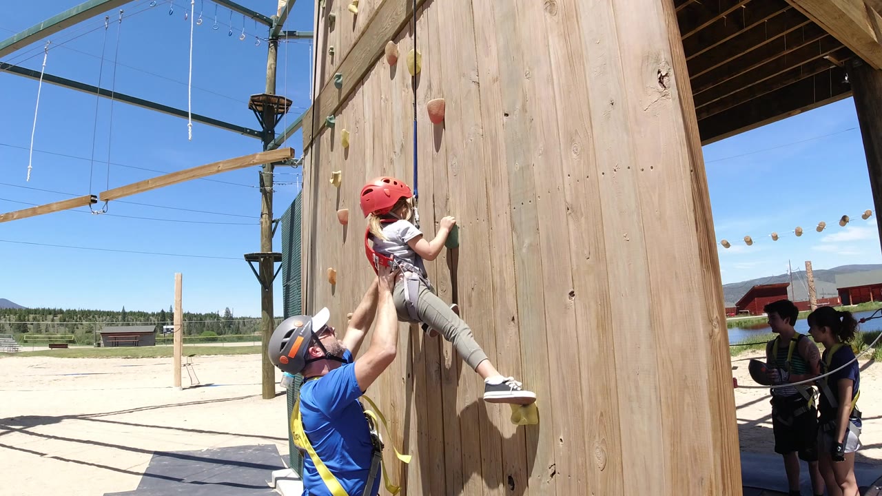One second in, the girl remains low on the wooden climbing wall as the adult lifts and guides her, with ropes overhead and other harnessed people waiting in the shaded area.