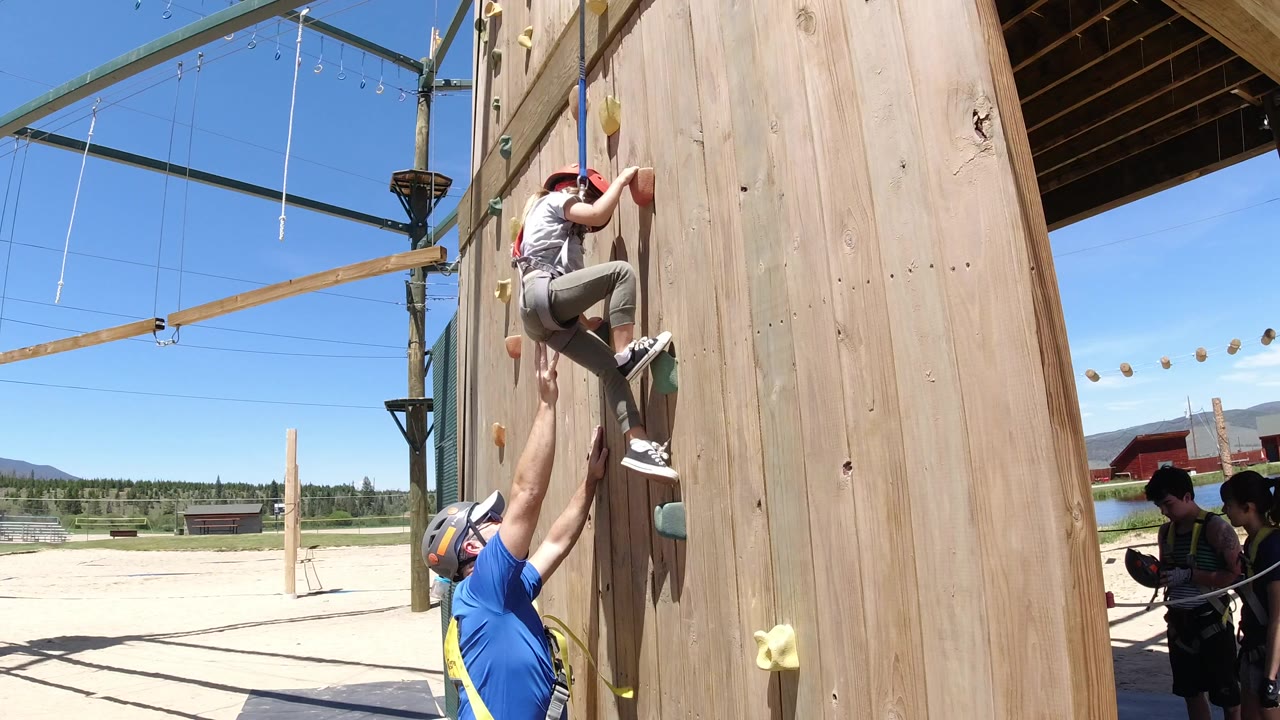 At Winter Park, Colorado, a boy in a red helmet climbs a wooden climbing wall while an adult in a blue shirt reaches up from below, with ropes-course lines and blue sky behind them.